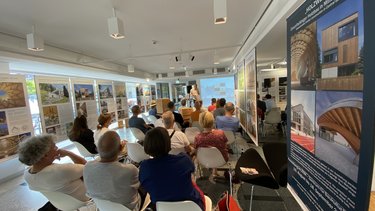 Publikum sitzt in einer Ausstellung und hört einen Vortrag zwischen Architekturtafeln zum Holzbau.