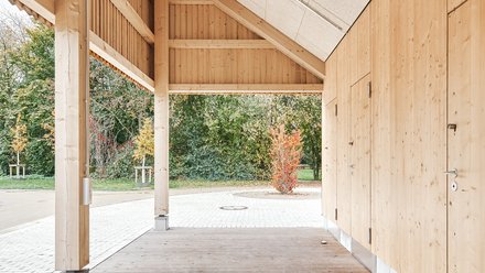 Überdachter Außenraum aus Holz mit sichtbaren Balken und hellen Deckenplatten; Blick auf Pflasterfläche und herbstliche Sträucher.