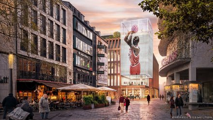 Stadtblick bei Dämmerung: Fußgängerzone mit Straßencafés links und rechts, im Zentrum ein neues, zurückgesetztes Gebäude mit großer, leuchtender Würfel‑Videofassade, die ein Basketballmotiv zeigt; Menschen flanieren, Bäume und historischere Nachbarbauten rahmen die Szene.