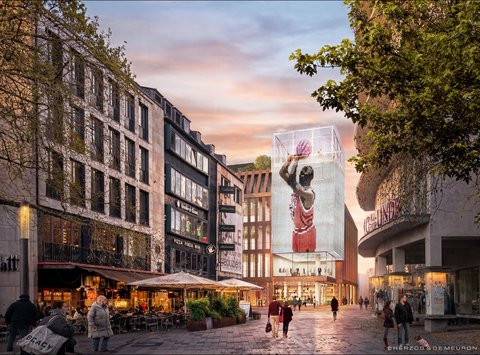 Stadtblick bei Dämmerung: Fußgängerzone mit Straßencafés links und rechts, im Zentrum ein neues, zurückgesetztes Gebäude mit großer, leuchtender Würfel‑Videofassade, die ein Basketballmotiv zeigt; Menschen flanieren, Bäume und historischere Nachbarbauten rahmen die Szene.