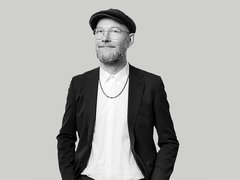 Schwarz‑weiß‑Porträt eines lächelnden Mannes mit Brille, kurzem Bart und Schiebermütze; er trägt ein weißes Hemd, eine Kette und ein dunkles Sakko, die Hände in den Taschen, vor neutralem hellgrauem Hintergrund. 