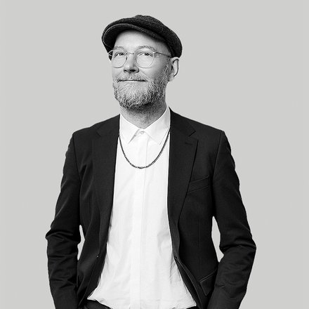 Schwarz‑weiß‑Porträt eines lächelnden Mannes mit Brille, kurzem Bart und Schiebermütze; er trägt ein weißes Hemd, eine Kette und ein dunkles Sakko, die Hände in den Taschen, vor neutralem hellgrauem Hintergrund. 