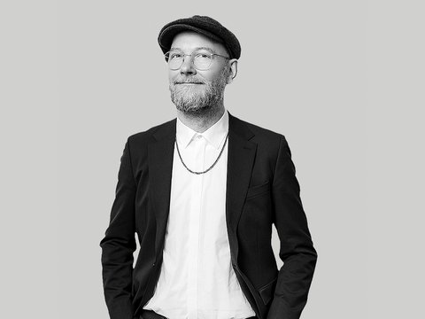 Schwarz‑weiß‑Porträt eines lächelnden Mannes mit Brille, kurzem Bart und Schiebermütze; er trägt ein weißes Hemd, eine Kette und ein dunkles Sakko, die Hände in den Taschen, vor neutralem hellgrauem Hintergrund. 