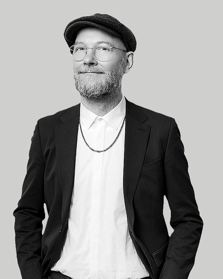 Schwarz‑weiß‑Porträt eines lächelnden Mannes mit Brille, kurzem Bart und Schiebermütze; er trägt ein weißes Hemd, eine Kette und ein dunkles Sakko, die Hände in den Taschen, vor neutralem hellgrauem Hintergrund. 