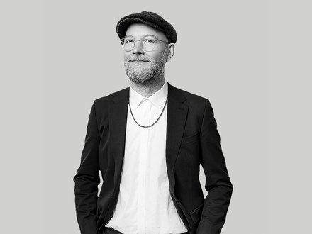 Schwarz‑weiß‑Porträt eines lächelnden Mannes mit Brille, kurzem Bart und Schiebermütze; er trägt ein weißes Hemd, eine Kette und ein dunkles Sakko, die Hände in den Taschen, vor neutralem hellgrauem Hintergrund. 