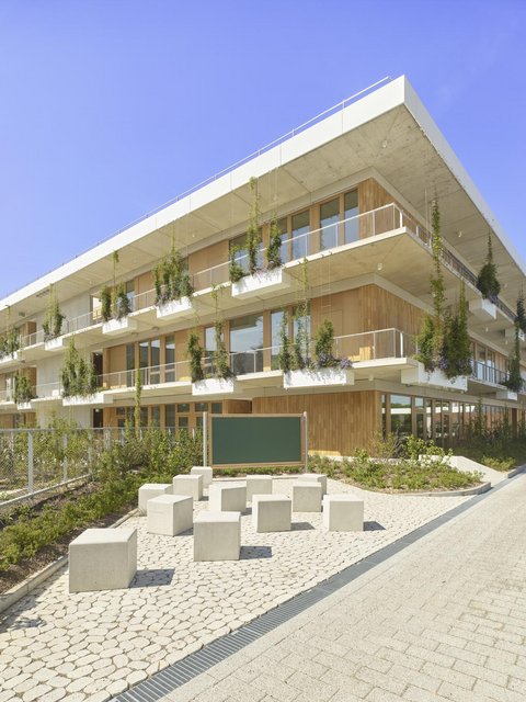Modernes dreistöckiges Gebäude mit Holzfassade und hängenden Pflanzen vor einem Sitzbereich mit quadratischen Betonhockern und einer grünen Tafel