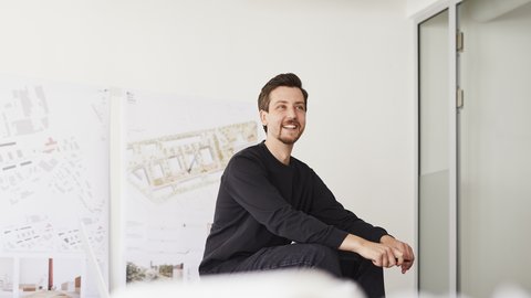Ein junger Mann sitzt entspannt in einem hellen Raum und lächelt. Hinter ihm stehen Tafeln oder Plakate mit Architekturzeichnungen. Er trägt dunkle Kleidung und wirkt, als sei er in ein Gespräch oder eine Präsentation vertieft.