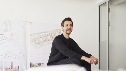 Ein junger Mann sitzt entspannt in einem hellen Raum und lächelt. Hinter ihm stehen Tafeln oder Plakate mit Architekturzeichnungen. Er trägt dunkle Kleidung und wirkt, als sei er in ein Gespräch oder eine Präsentation vertieft.