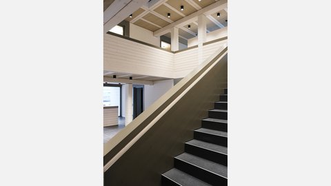 Blick entlang eines breiten Treppenlaufs zur Galerie mit Holzbrüstung; Rasterdecke mit sichtbaren Balken und Spotleuchten.