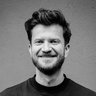 "Porträt eines lächelnden Mannes mit vollem dunklem Bart, stylishem Haarschnitt und schwarzem T-Shirt vor grauer Betonwand. Der Mann blickt direkt in die Kamera."