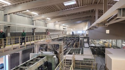 Innenansicht einer Holzindustriehalle: Laufstege über komplexen Förder‑ und Sägelinien, wenige Personen auf dem Steg, hohe Decke.