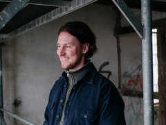 Halbporträt einer lächelnden Person, die nach links blickt; dunkle Jacke über Rollkragenpullover, Hintergrund mit Gerüst und rohem Beton in einem Industrie-/Bauraum.