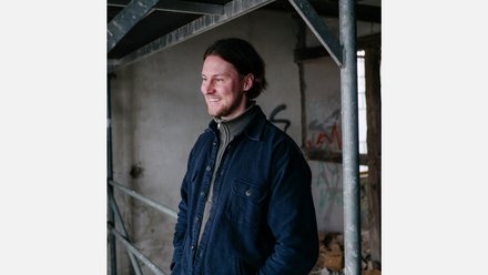 Halbporträt einer lächelnden Person, die nach links blickt; dunkle Jacke über Rollkragenpullover, Hintergrund mit Gerüst und rohem Beton in einem Industrie-/Bauraum.