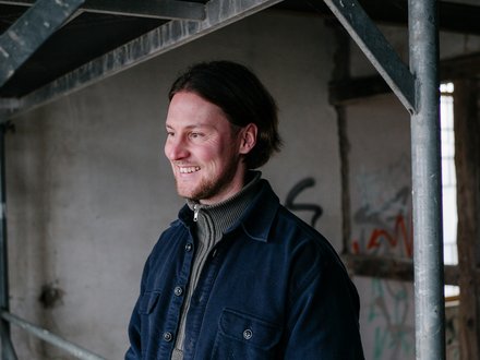 Halbporträt einer lächelnden Person, die nach links blickt; dunkle Jacke über Rollkragenpullover, Hintergrund mit Gerüst und rohem Beton in einem Industrie-/Bauraum.