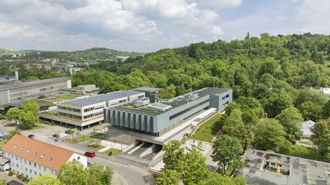 Luftaufnahme eines modernen Campusgebäudes mit umliegenden Bäumen und weiteren Gebäuden in einer hügeligen Landschaft