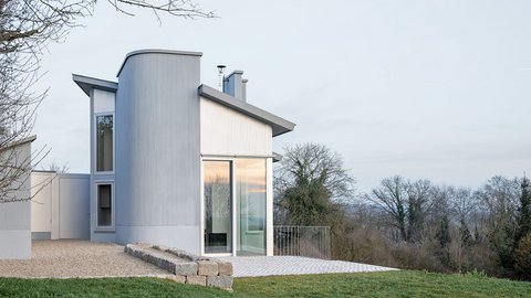 Modernes Ferienhaus mit großen Fenstern und abgerundetem Anbau auf einem leicht abfallenden Grundstück mit Rasen und Bäumen im Hintergrund