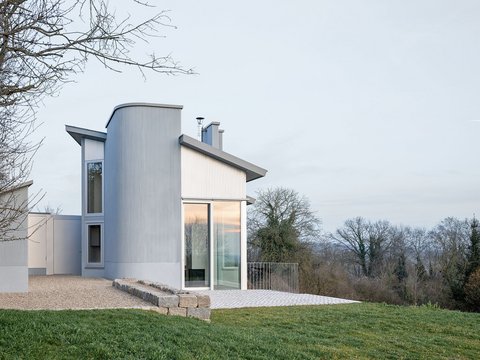 Modernes Ferienhaus mit großen Fenstern und abgerundetem Anbau auf einem leicht abfallenden Grundstück mit Rasen und Bäumen im Hintergrund