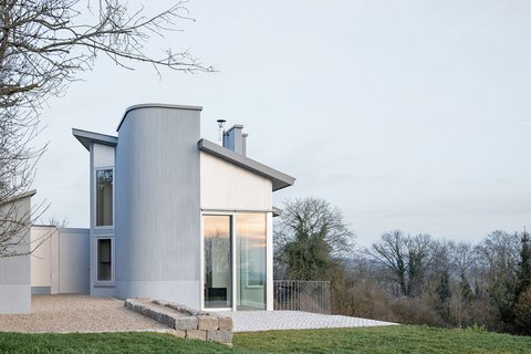 Modernes Ferienhaus mit großen Fenstern und abgerundetem Anbau auf einem leicht abfallenden Grundstück mit Rasen und Bäumen im Hintergrund