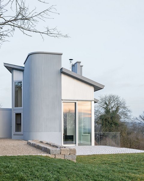 Modernes Ferienhaus mit großen Fenstern und abgerundetem Anbau auf einem leicht abfallenden Grundstück mit Rasen und Bäumen im Hintergrund