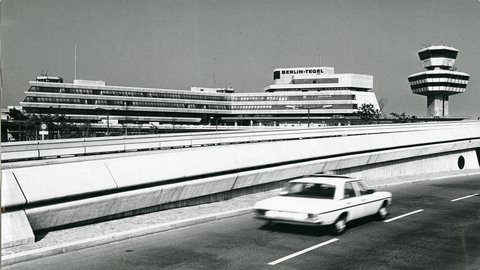 Eine historische Schwarz-Weiß-Fotografie aus einer tiefen Perspektive zeigt das Hauptgebäude und den Kontrollturm des Flughafens Berlin-Tegel. Das Terminal ist ein langes, gestuftes Gebäude aus Beton und Glas mit dem Schriftzug "BERLIN-TEGEL" auf dem Dach. Rechts davon steht der markante, polygonale Kontrollturm auf einem schlanken Betonpfeiler. Im Vordergrund fährt ein weißes Auto, ein Mercedes-Benz aus den 1970er Jahren, auf einer mehrspurigen Straße, davor verläuft eine erhöhte Betonstruktur. Die Szene wirkt ruhig und modern für die Zeit.