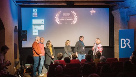 Fünf Personen stehen auf einer Bühne vor einer großen Leinwand. Die Leinwand zeigt ein Festival-Logo mit Lorbeerkranz und Text "Architekturreis 2024". Links sitzt ein Publikum auf roten Samtsesseln. Blauviolette Beleuchtung und ein BR-Logo auf der rechten Seite.