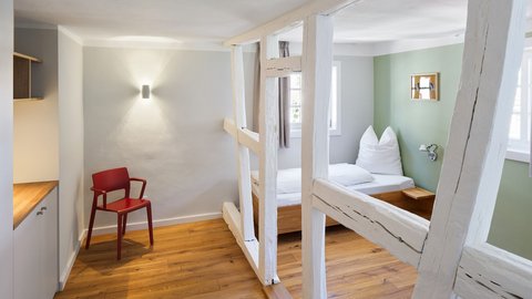 "Modernes Schlafzimmer mit offener Raumaufteilung. Links rote Holzstühle auf hellem Holzboden. Mitte und rechts von weißen Holzpfosten abgeteilter Schlafbereich mit grüner Wandfarbe. Einzelnes Bett mit weißer Bettwäsche und braunem Nachttisch. Hellgrüne Wand, mehrere Fenster mit Tageslicht, minimalistische Einrichtung."