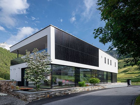 „Ein modernes, zweistöckiges Gebäude mit verglastem Erdgeschoss und fassadenintegrierten Photovoltaik-Modulen am weißen Obergeschoss vor einer grünen Hügellandschaft.“