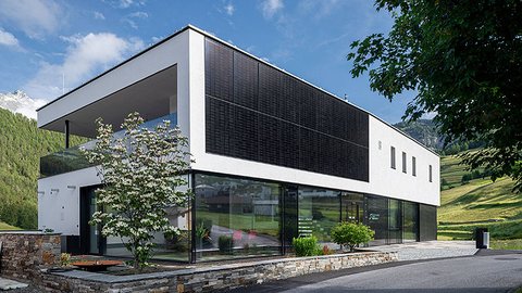 „Ein modernes, zweistöckiges Gebäude mit verglastem Erdgeschoss und fassadenintegrierten Photovoltaik-Modulen am weißen Obergeschoss vor einer grünen Hügellandschaft.“