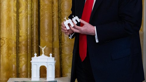 Nahaufnahme einer Person in einem dunklen Anzug und einer roten Krawatte. Die Person hält zwei kleine, weiße Architekturmodelle eines Triumphbogens in den Händen. Ein größeres, gleichartiges Modell mit einer goldenen, geflügelten Figur an der Spitze steht im Vordergrund auf einer Oberfläche. Im Hintergrund hängt ein goldfarbener Vorhang mit Muster.