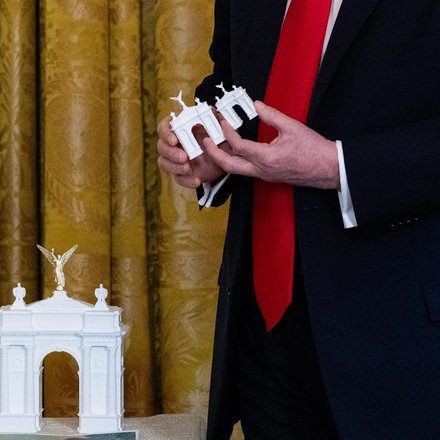 Nahaufnahme einer Person in einem dunklen Anzug und einer roten Krawatte. Die Person hält zwei kleine, weiße Architekturmodelle eines Triumphbogens in den Händen. Ein größeres, gleichartiges Modell mit einer goldenen, geflügelten Figur an der Spitze steht im Vordergrund auf einer Oberfläche. Im Hintergrund hängt ein goldfarbener Vorhang mit Muster.