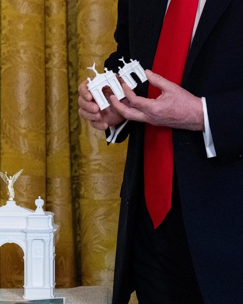 Nahaufnahme einer Person in einem dunklen Anzug und einer roten Krawatte. Die Person hält zwei kleine, weiße Architekturmodelle eines Triumphbogens in den Händen. Ein größeres, gleichartiges Modell mit einer goldenen, geflügelten Figur an der Spitze steht im Vordergrund auf einer Oberfläche. Im Hintergrund hängt ein goldfarbener Vorhang mit Muster.