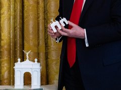 Nahaufnahme einer Person in einem dunklen Anzug und einer roten Krawatte. Die Person hält zwei kleine, weiße Architekturmodelle eines Triumphbogens in den Händen. Ein größeres, gleichartiges Modell mit einer goldenen, geflügelten Figur an der Spitze steht im Vordergrund auf einer Oberfläche. Im Hintergrund hängt ein goldfarbener Vorhang mit Muster.