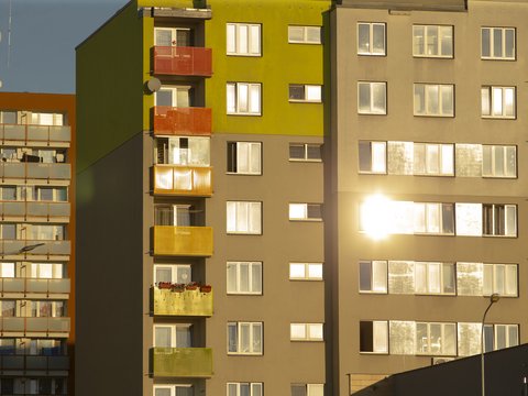 "Moderne Wohnhäuser an sonnigem Tag. Im Fokus steht ein Gebäude mit gelb-grüner Fassade und farbigen Balkonen in Orange, Rot und Gelb. Daneben ein beige-graues Gebäude. Mehrere Fenster reflektieren das Sonnenlicht. Links teilweise ein älteres Gebäude mit brauner Fassade sichtbar."