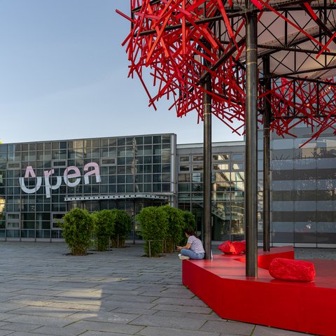 Rote, abstrakte Skulptur aus vielen Stäben auf rotem Sockel vor einem modernen Glasgebäude mit der Aufschrift 'Opera'