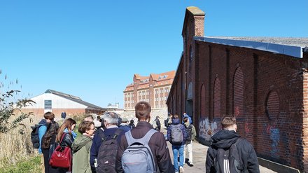 Gruppe von Menschen geht über ein ehemaliges Industriegelände entlang eines Backsteingebäudes.