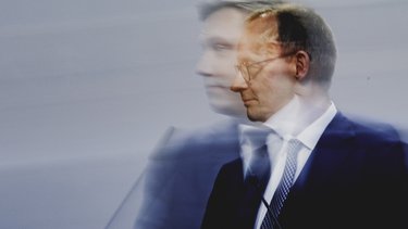 Pressefoto eines Politikers am Rednerpult, Seitenansicht mit Bewegungsunschärfe vor neutralem Hintergrund.