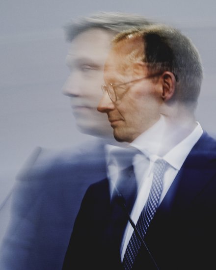 Pressefoto eines Politikers am Rednerpult, Seitenansicht mit Bewegungsunschärfe vor neutralem Hintergrund.