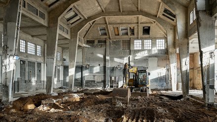 "Großer Betonbau in Ruinen mit freigelegtem Dach. Ein gelber Bagger bewegt Schuttmaterial im Inneren. Massive Betonsäulen und -träger sind sichtbar. Kaputte Fenster, teils offene Mauern. Naturlicht fällt durch mehrere Öffnungen herein."
