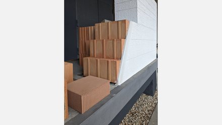 Detail Wandaufbau: gestufte, gerillte Ziegel hinter weiß verputzter Außenfassade auf Betonsockel; daneben Kiesbett.