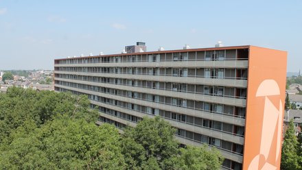 Langer Wohnriegel mit durchgehenden Balkonen; orangefarbener Stirngiebel mit großer Grafik, Baumkronen im Vordergrund und klarer Himmel.