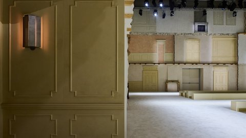 "Ausstellungsraum mit Architektur-Installation aus übergroßen historischen Architektur-Elementen. Die Wand auf der linken Seite zeigt eine olivgelbe Panelstruktur mit integrierter Wandleuchte. Die rechte Seite präsentiert mehrere klassische Türen in verschiedenen Farben und Materialien (Creme, Grün, Rosa, Marmor) sowie Fensterelemente. Eine schwarze Vintage-Kaminumrandung steht zentriert. Eine Treppe mit Holzbrüstung verbindet mehrere Ebenen. Der Konkreetboden verstärkt den skulpturalen Charakter."