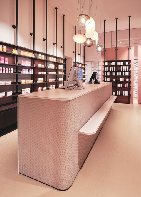 „Ein modernes, komplett in Pastellpink gestaltetes Geschäft. Im Vordergrund steht ein langer, abgerundeter Tresen, dahinter befinden sich dunkle, gut gefüllte Regale und dekorative Hängelampen.“