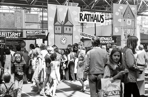 "Historische Schwarzweiß-Aufnahme einer belebten Menschenmenge an einem Bahnhof mit Schild 'RATHAUS'. Ein großes Schild zeigt eine Kirche mit zwei Türmen und eine Uhr. Männer, Frauen und Kinder verschiedener Altersgruppen stehen unter Bahnhofsdächern. Links sind Schilder mit 'ARBEITSAMT' und 'FORMATION' sichtbar."