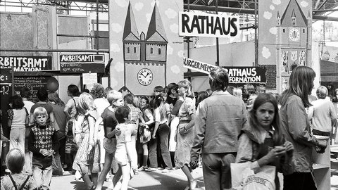 "Historische Schwarzweiß-Aufnahme einer belebten Menschenmenge an einem Bahnhof mit Schild 'RATHAUS'. Ein großes Schild zeigt eine Kirche mit zwei Türmen und eine Uhr. Männer, Frauen und Kinder verschiedener Altersgruppen stehen unter Bahnhofsdächern. Links sind Schilder mit 'ARBEITSAMT' und 'FORMATION' sichtbar."