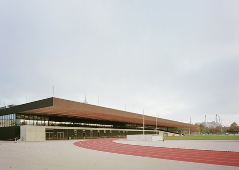 Campusgebäude mit weit auskragendem Dach und Laufbahn im Vordergrund.