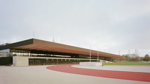 Campusgebäude mit weit auskragendem Dach und Laufbahn im Vordergrund.