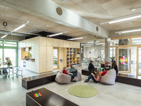 Offener Schul- und Aufenthaltsraum mit Holzeinbauten, Tischen und Sitzsäcken. Kinder lesen und unterhalten sich in kleinen Gruppen.