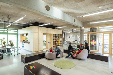 Offener Schul- und Aufenthaltsraum mit Holzeinbauten, Tischen und Sitzsäcken. Kinder lesen und unterhalten sich in kleinen Gruppen.
