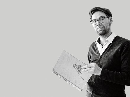 Schwarz-weiß-Porträt: Eine Person mit Brille und Mütze steht halber Oberkörper nach rechts im Bild und hält ein großes Buch mit Illustration auf dem Einband. Viel leere, hellgraue Fläche links