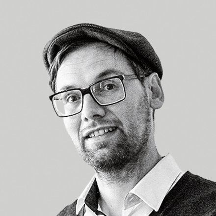 Schwarz-weiß-Porträt: Eine Person mit Brille und Mütze steht halber Oberkörper nach rechts im Bild und hält ein großes Buch mit Illustration auf dem Einband. Viel leere, hellgraue Fläche links